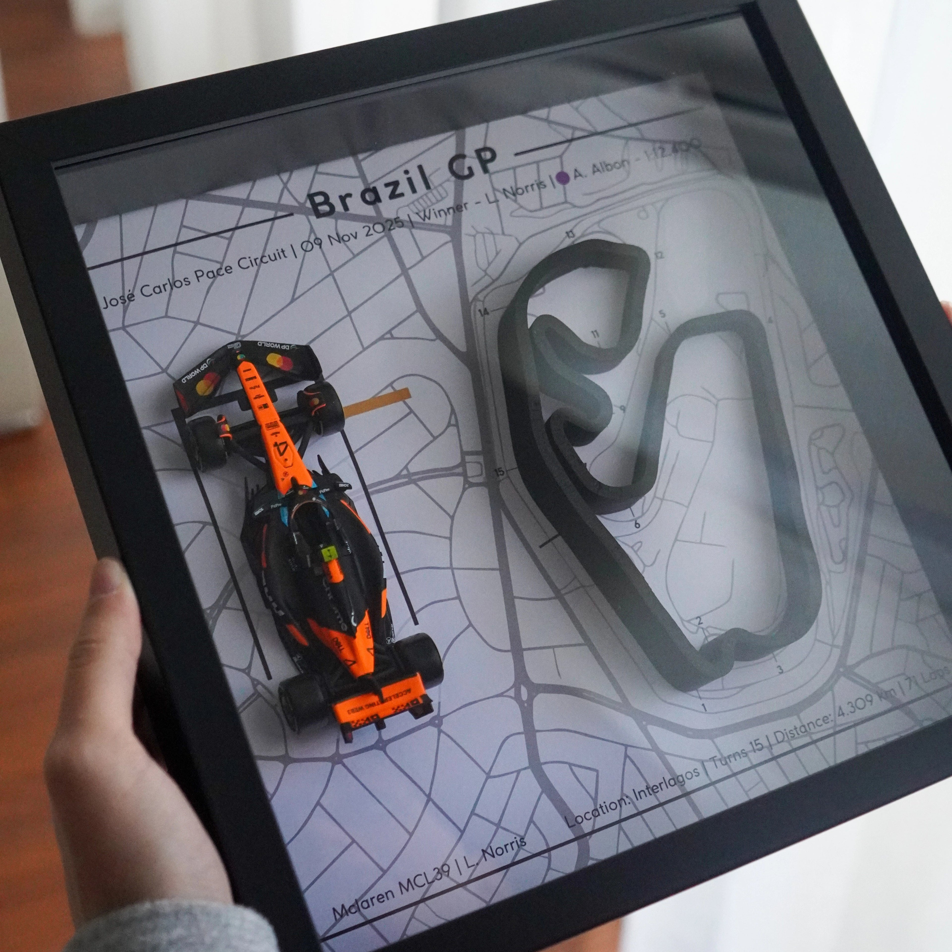 Framed artwork with a racing car and track layout on a map, held by a person.